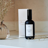 Black bottle with white label on a white surface next to a vase with dried flowers and a box.