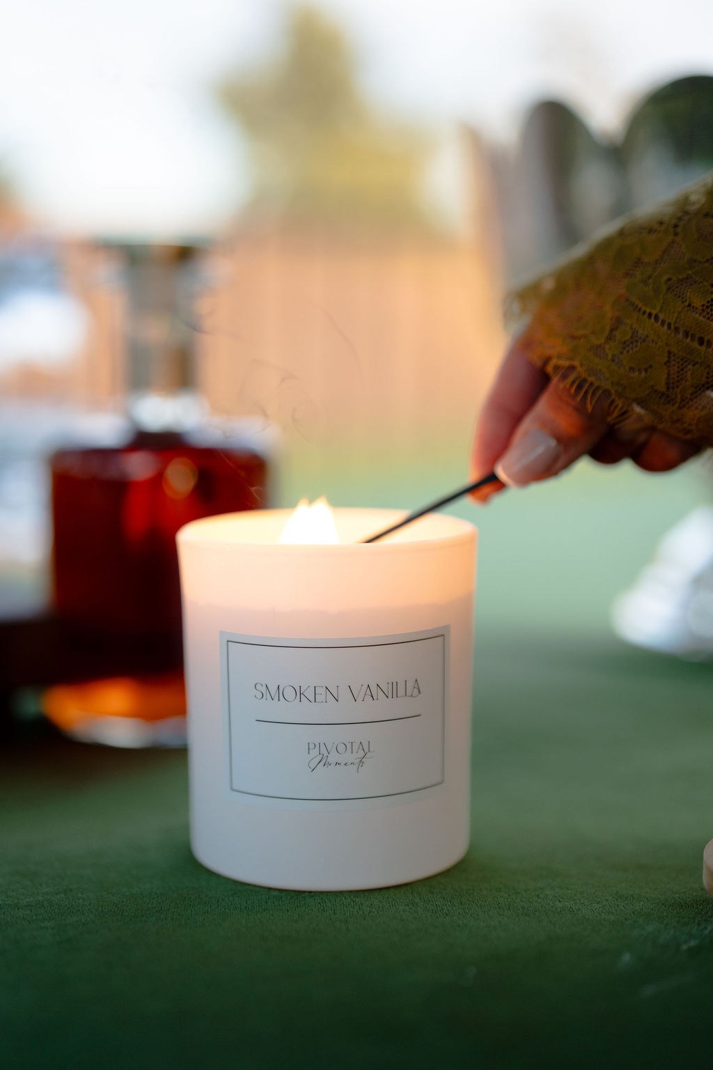 Hand lighting a Pivotal Moments Smoken Vanilla candle in a white glass vessel with a long match, a bourbon decanter glowing softly in the background.