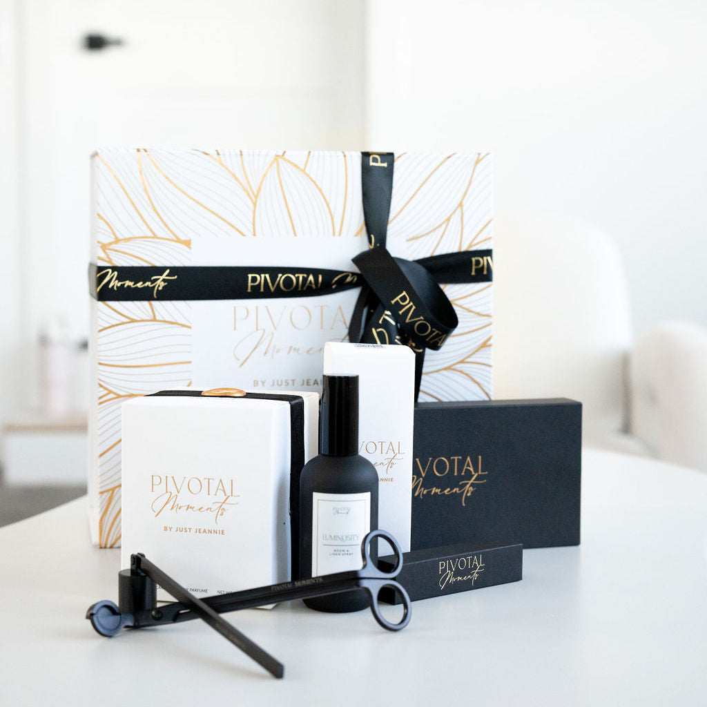 A gift set from Pivotal Moments including a candle and room spray with gold-tipped containers, accompanied by custom gold-tipped matches and a candle care set, all displayed on a white surface.