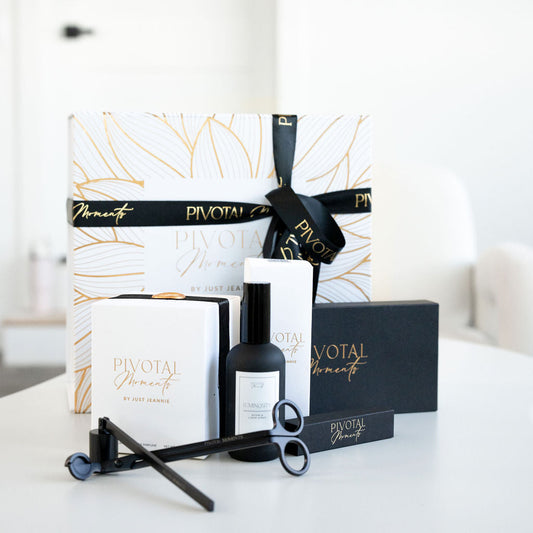 A gift set from Pivotal Moments including a candle and room spray with gold-tipped containers, accompanied by custom gold-tipped matches and a candle care set, all displayed on a white surface.