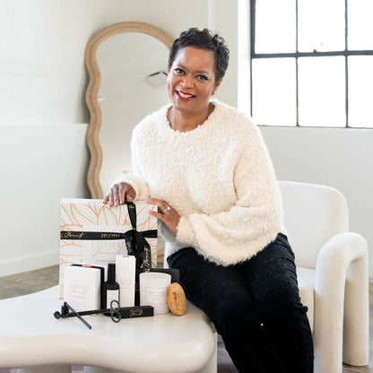 A woman sitting on a modern white bench in a room with a mirror and a window, holding a luxurious gift box with a 'Warm & Cozy' label.