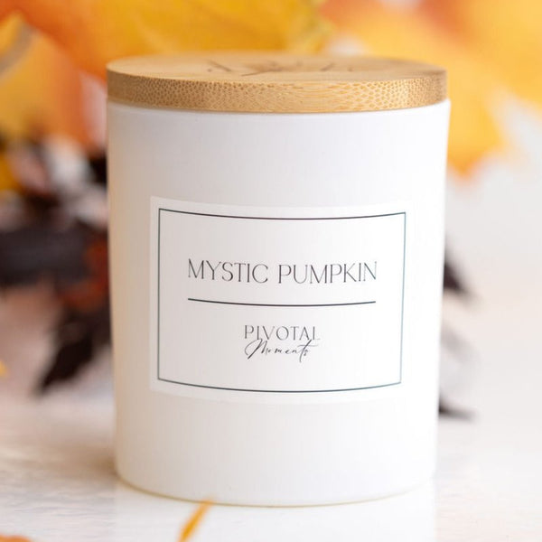 A white cylindrical glass candle with a wooden lid, labeled 'MYSTERY PUMPKIN' and 'Pivotal Moments'. The candle is placed against a white background.