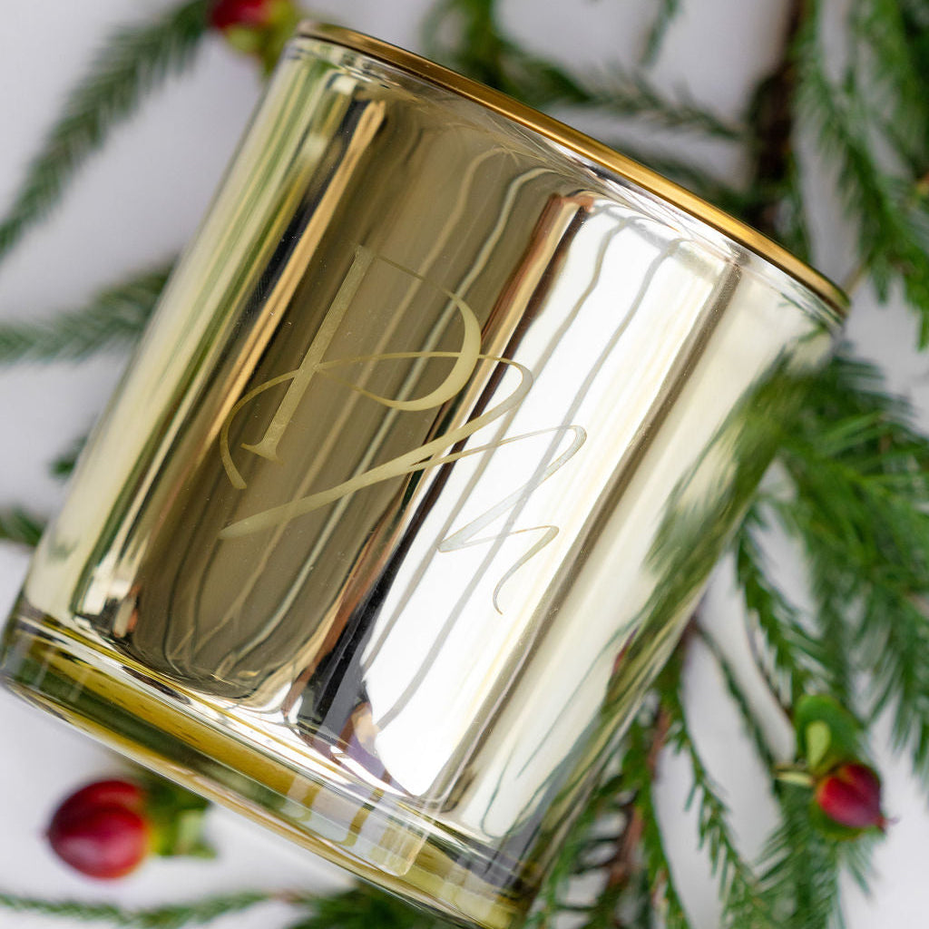 A clear glass candle with a gold-colored lid, featuring the brand logo 'PM' on the front.