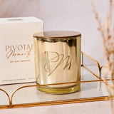 Gold candle with 'PM' logo on a glass shelf with a white box in the background