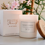 Pivotal Moments candle with packaging and wooden lid on a surface with flowers in the background