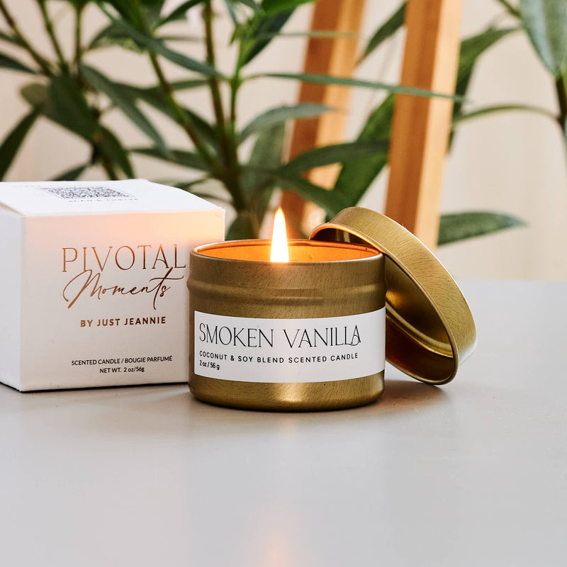 Smoked Vanilla candle with a lit flame on a table next to a plant and box.