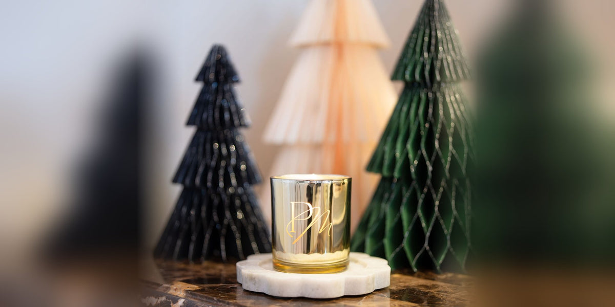 Gold glass Pivotal Moments Christmas candle with PM monogram on a marble tray among black, blush and green paper Christmas trees