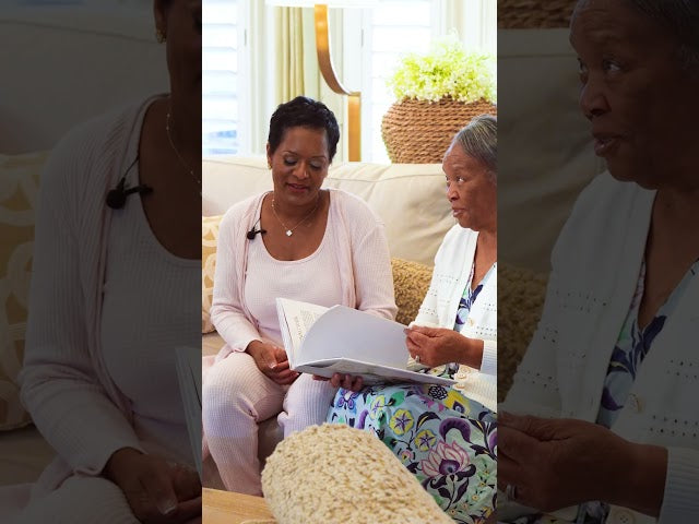 Two women sitting on a couch, one holding a book, in a home setting.